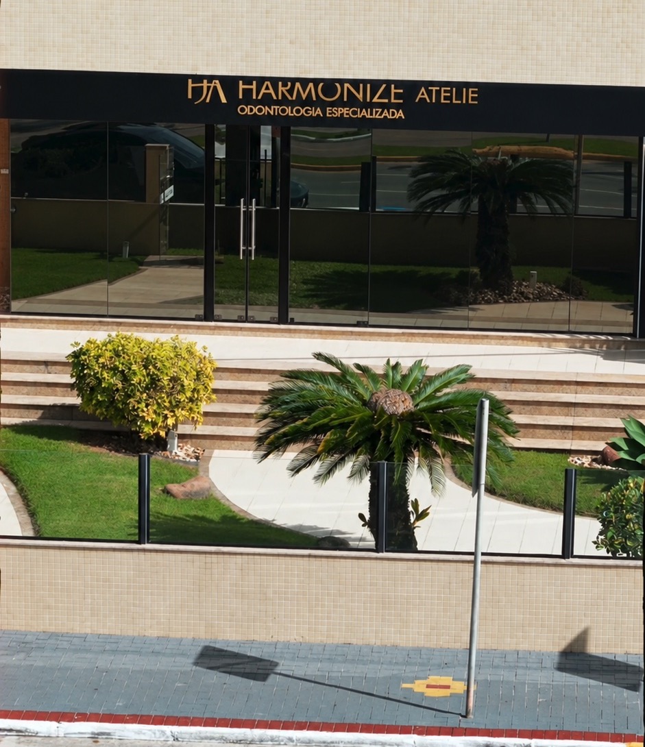 Fachada da Harmonize Ateliê sem pessoas, com letreiro dourado, entrada envidraçada e jardim; imagem editada a partir de fotografia real